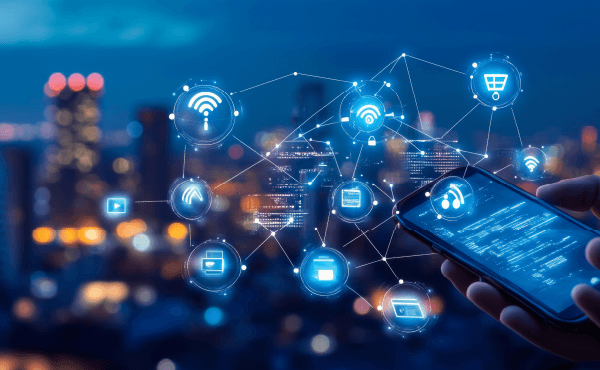 Na zdjęciu smartfon w dłoni, nad którym unosi się cyfrowa siatka z ikonkami symbolizującymi urządzenia i usługi cyfrowe: sieć wi-fi, kartę płatniczą, ekran urządzenia. W tle rozświetlone wieżowce.